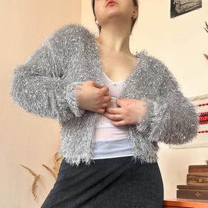 Könnte beinhalten: Ein silberfarbener, flauschiger Cardigan mit offenem Schnitt. Der Cardigan hat einen V-Ausschnitt und lange Ärmel. Das Material scheint ein weiches, strukturiertes Garn mit leichtem Glanz zu sein. Der Cardigan wird über einem weißen Oberteil und mit einem dunklen Rock getragen.
