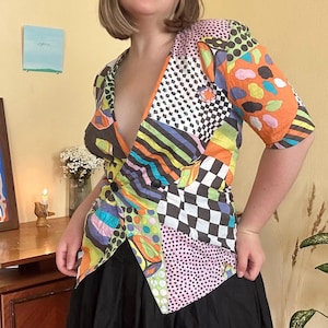May include: A short-sleeved blazer featuring a vibrant patchwork design with various patterns including black and white checkers, polka dots, stripes, and abstract shapes in orange, green, purple, and blue. The blazer has a deep V-neckline and two black buttons.