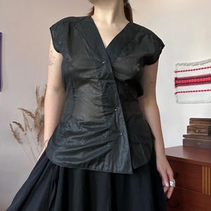 May include: A black sleeveless blouse featuring a V-neckline and an asymmetrical front closure with four visible buttons. The garment is made of a semi-sheer, lightweight fabric with subtle vertical paneling and a slightly curved hemline.