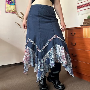 May include: A navy blue midi skirt featuring a textured fabric, a V-shaped ruffled seam across the mid-section, and a multi-colored floral patterned hemline with an asymmetrical handkerchief cut that falls below the knee in a layered design.