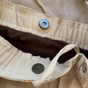 May include: A beige woven fabric bag featuring a dark brown interior lining with a zippered pocket. The bag includes a magnetic snap closure, a drawstring cord, and small circular mirror embellishments on the exterior surface.