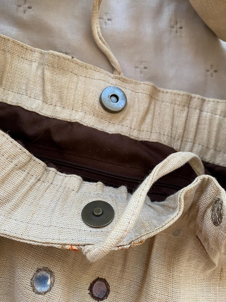 May include: A beige woven fabric bag featuring a dark brown interior lining with a zippered pocket. The bag includes a magnetic snap closure, a drawstring cord, and small circular mirror embellishments on the exterior surface.