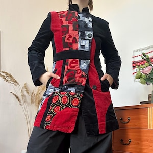 May include: A long-sleeved jacket featuring a black base with a patchwork front panel. The front panel displays red, grey, and black sections with abstract patterns, including circular motifs and letters. It has a high collar and three buttons.