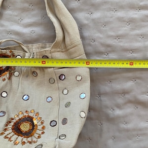 May include: A beige fabric shoulder bag decorated with small circular mirrors and a large floral-style sequin embroidery. A yellow measuring tape is placed across the bag, showing a width of approximately 15 inches from the center to the edge.