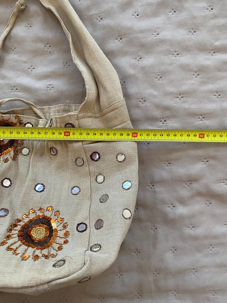 May include: A beige fabric shoulder bag decorated with small circular mirrors and a large floral-style sequin embroidery. A yellow measuring tape is placed across the bag, showing a width of approximately 15 inches from the center to the edge.