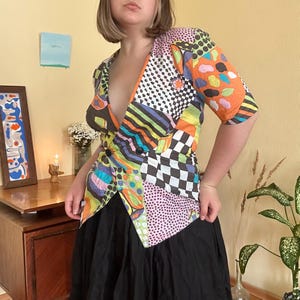 Pode incluir: Um blazer colorido em patchwork com um padrão geométrico arrojado em várias cores, incluindo laranja, preto, branco e rosa. O blazer tem mangas curtas e um design de abotoamento duplo. A modelo está usando uma saia preta.