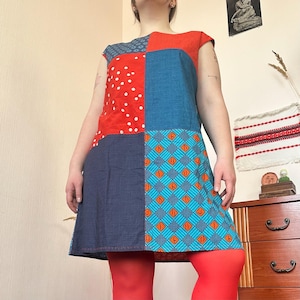 May include: A patchwork dress with a vibrant design. The dress features squares of red with white polka dots, blue, and patterned fabric in blue and orange. The dress has short sleeves and is paired with red tights.