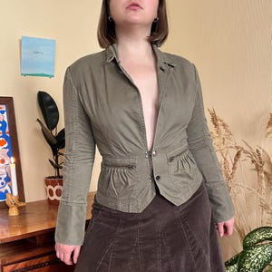 May include: An olive green jacket with a tailored fit, featuring a notched collar, two front pockets with zippers, and a button closure. The jacket is paired with a brown corduroy skirt. The overall style is casual and chic.