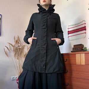 May include: A black long-sleeve coat featuring a high collar with a drawstring, a central vertical panel with gathered fabric detailing, and two side pockets. The coat has a smooth finish and a structured fit that extends to the mid-thigh area.