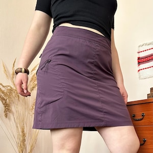 以下が含まれることがあります： 左側にジッパーポケットが付いたプラム色のスカート。スカートはストレートで、わずかにAラインカットです。モデルは黒いトップスと木製のブレスレットを着用しています。スカートは軽量素材で作られています。