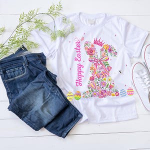 Puede incluir: Camiseta blanca con un diseño de conejito floral, una corona rosa y la frase "Happy Easter". El conejito está hecho de flores rosas, amarillas y moradas. También hay unos vaqueros azules y zapatillas blancas en la imagen.