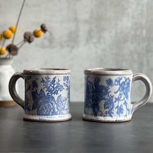 May include: Two ceramic mugs with blue floral patterns on a white background. The mugs have a rectangular shape with a handle and a brown rim. The mugs are placed on a dark surface.