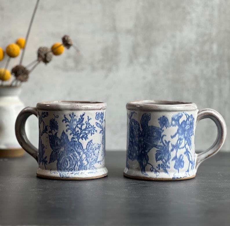 May include: Two ceramic mugs with blue floral patterns on a white background. The mugs have a rectangular shape with a handle and a brown rim. The mugs are placed on a dark surface.