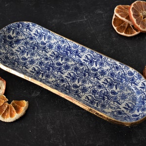 May include: A white ceramic serving dish with a blue and gold floral pattern. The dish is oval shaped and has a gold rim.