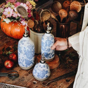 May include: Three ceramic oil dispensers featuring a blue and white floral pattern with silver-toned metal pour spouts. The set includes two tall cylindrical bottles and one shorter, rounded bottle, all with a natural beige base finish.