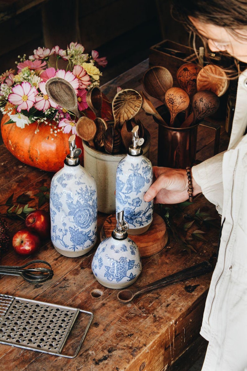 May include: Three ceramic oil dispensers featuring a blue and white floral pattern with silver-toned metal pour spouts. The set includes two tall cylindrical bottles and one shorter, rounded bottle, all with a natural beige base finish.