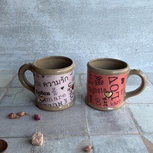 Pair of violet and coral pottery mugs featuring words for love in various languages, 24k gold accents, and romantic props like dried roses and sugar, ideal for galentines or valentines gifting