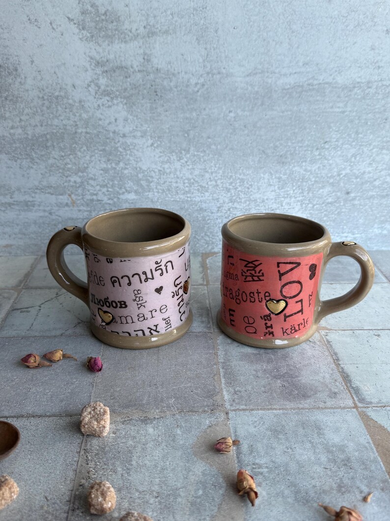 Pair of violet and coral pottery mugs featuring words for love in various languages, 24k gold accents, and romantic props like dried roses and sugar, ideal for galentines or valentines gifting