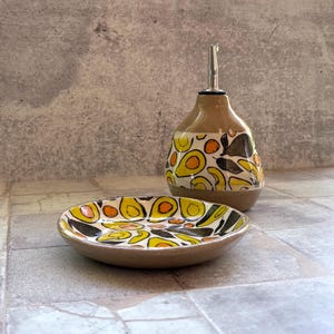 May include: A ceramic oil bottle and small dish set. The bottle has a brown base and a patterned design of yellow, orange, and black avocado shapes. The dish mirrors the design. The bottle has a silver spout.