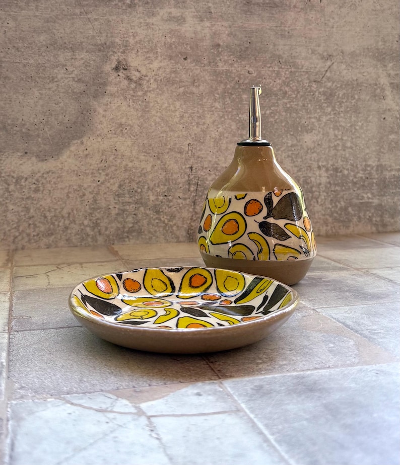 May include: A ceramic oil bottle and small dish set. The bottle has a brown base and a patterned design of yellow, orange, and black avocado shapes. The dish mirrors the design. The bottle has a silver spout.