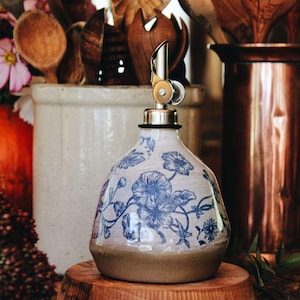 May include: A ceramic oil dispenser with a rounded base and a tapered neck, featuring a blue floral pattern on a cream-colored background. The dispenser is topped with a silver-colored metal pour spout with a hinged lid mechanism.