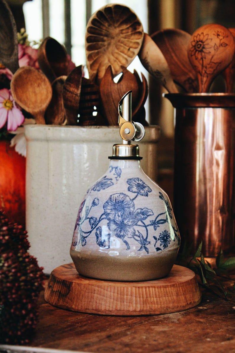 May include: A ceramic oil dispenser with a rounded base and a tapered neck, featuring a blue floral pattern on a cream-colored background. The dispenser is topped with a silver-colored metal pour spout with a hinged lid mechanism.