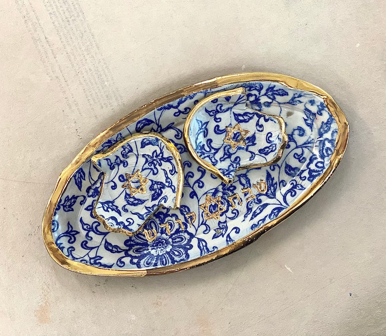Op de afbeelding: Een keramische serveerset met een blauw-wit bloemmotief en gouden rand. De set bevat een ovale schaal en twee kleinere schaaltjes, elk met een gouden Davidster. Er is Hebreeuwse tekst zichtbaar.