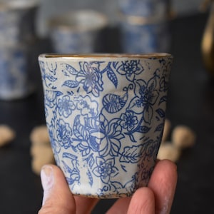 May include: A white ceramic cup with a blue floral pattern and a gold rim. The cup is being held in a hand.