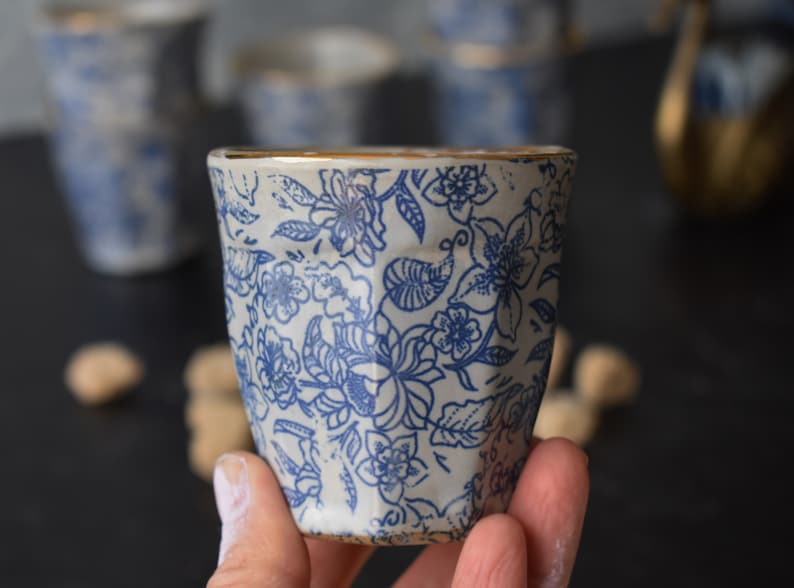 May include: A white ceramic cup with a blue floral pattern and a gold rim. The cup is being held in a hand.