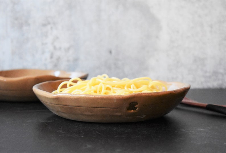 Puede incluir: Primer plano de un cuenco de cer&aacute;mica marr&oacute;n lleno de fideos espagueti cocidos. El cuenco tiene un aspecto r&uacute;stico y hecho a mano y est&aacute; sobre una superficie negra.