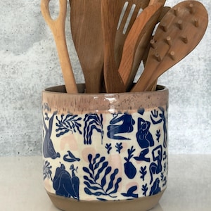 May include: A cylindrical ceramic utensil holder with a cream-coloured base featuring a blue and light pink abstract pattern of human figures and foliage. The container is filled with various wooden kitchen utensils, including spoons and a pasta server.