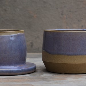 Peut inclure: Deux pi&egrave;ces en c&eacute;ramique faites &agrave; la main. L'une est un beurrier avec un couvercle, un gla&ccedil;age lavande et un bord beige. L'autre est un r&eacute;cipient cylindrique assorti avec une base et un bord beiges. Les deux sont sur une surface en bois.