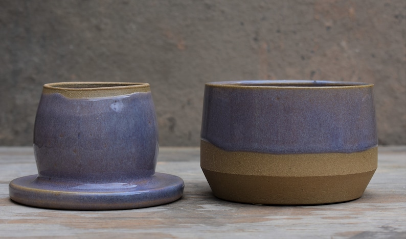 Peut inclure: Deux pi&egrave;ces en c&eacute;ramique faites &agrave; la main. L'une est un beurrier avec un couvercle, un gla&ccedil;age lavande et un bord beige. L'autre est un r&eacute;cipient cylindrique assorti avec une base et un bord beiges. Les deux sont sur une surface en bois.