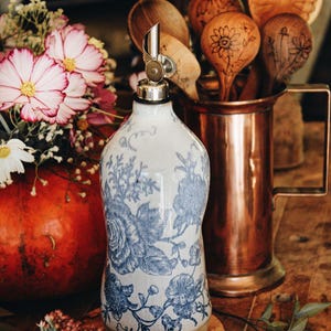 May include: A ceramic oil dispenser bottle featuring a white base with intricate blue floral patterns and a metallic pour spout. The bottle has a rounded body and a flat base, showcasing a detailed botanical design in a classic blue and white color scheme.