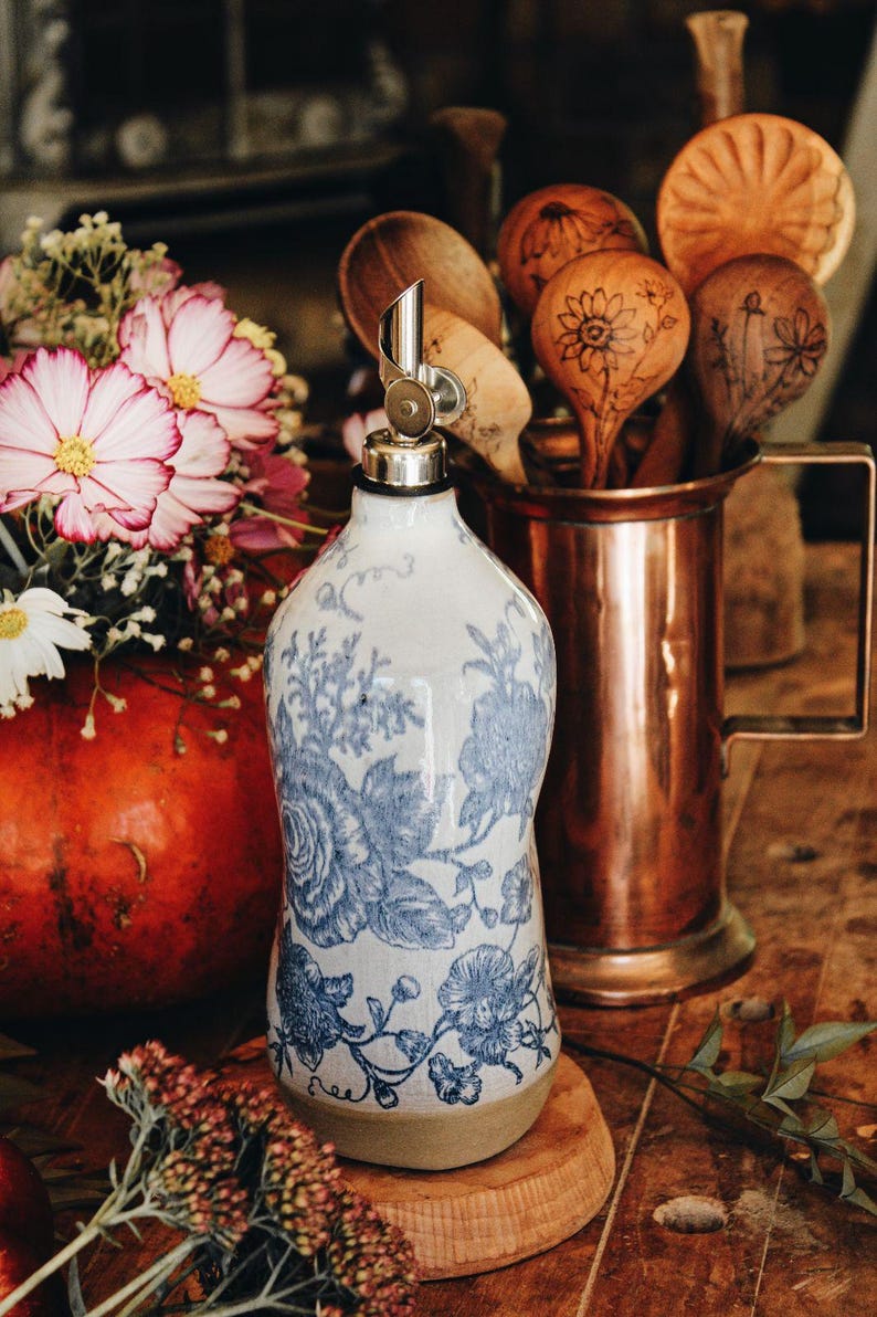 May include: A ceramic oil dispenser bottle featuring a white base with intricate blue floral patterns and a metallic pour spout. The bottle has a rounded body and a flat base, showcasing a detailed botanical design in a classic blue and white color scheme.