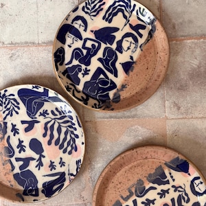 May include: Three ceramic plates with a cream-colored background, featuring navy blue illustrations of figures and foliage, and pink accents. The plates have a speckled, light brown rim.