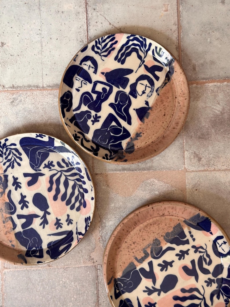May include: Three ceramic plates with a cream-colored background, featuring navy blue illustrations of figures and foliage, and pink accents. The plates have a speckled, light brown rim.