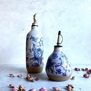 May include: Two ceramic oil dispensers with a white glaze and blue floral pattern. The taller bottle is cylindrical with a metal pour spout, while the shorter bottle is bulbous with a black cap. Both have an unglazed brown base.