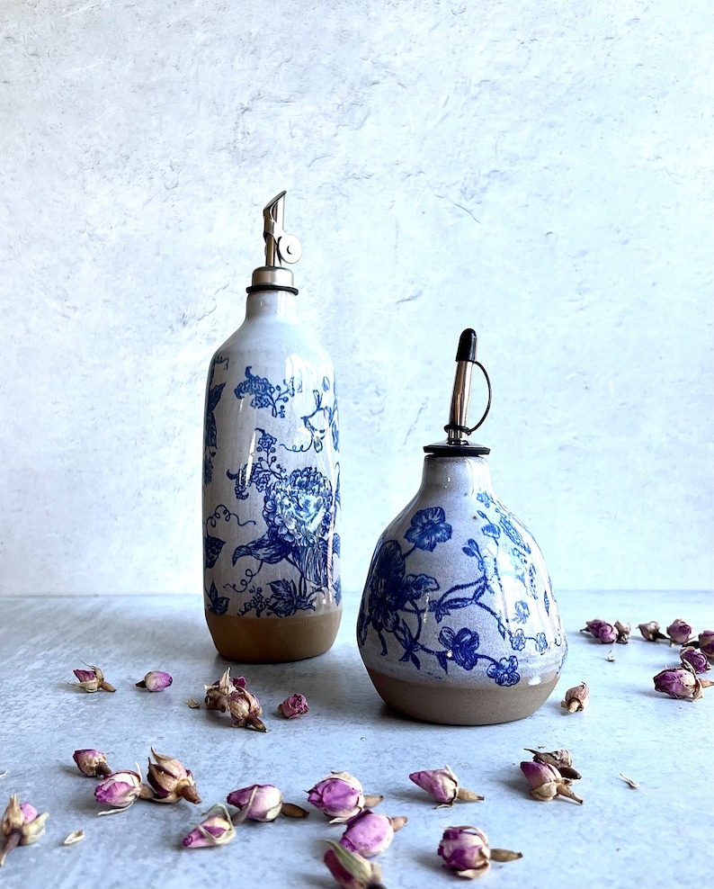 May include: Two ceramic oil dispensers with a white glaze and blue floral pattern. The taller bottle is cylindrical with a metal pour spout, while the shorter bottle is bulbous with a black cap. Both have an unglazed brown base.