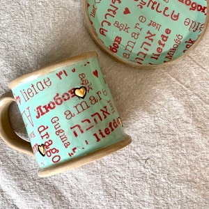 May include: A ceramic mug and saucer set. The mug and saucer are light teal with red text in various languages, including "love" and "amore." Gold heart accents are on the mug. The rim and handle are beige.