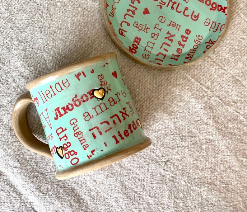 May include: A ceramic mug and saucer set. The mug and saucer are light teal with red text in various languages, including "love" and "amore." Gold heart accents are on the mug. The rim and handle are beige.