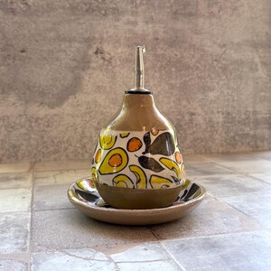 May include: A ceramic olive oil dispenser with a stainless steel spout, resting on a matching saucer. The dispenser is tan with a colorful pattern of yellow, black, and green avocado and leaf designs.