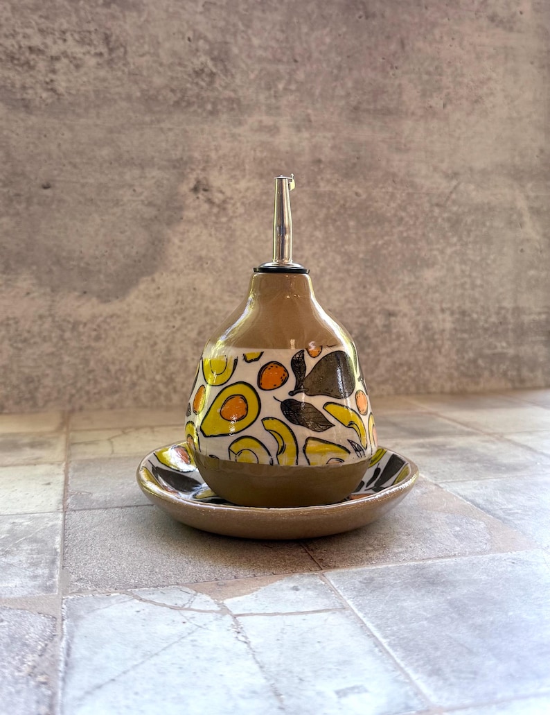 May include: A ceramic olive oil dispenser with a stainless steel spout, resting on a matching saucer. The dispenser is tan with a colorful pattern of yellow, black, and green avocado and leaf designs.