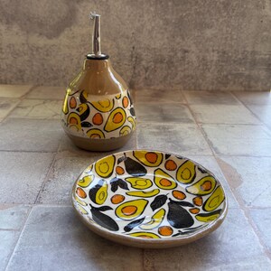 May include: A ceramic oil bottle and small dish set. The bottle has a brown base and a silver spout, decorated with yellow, orange, and black avocado designs. The matching dish features the same avocado pattern. A unique kitchenware set.