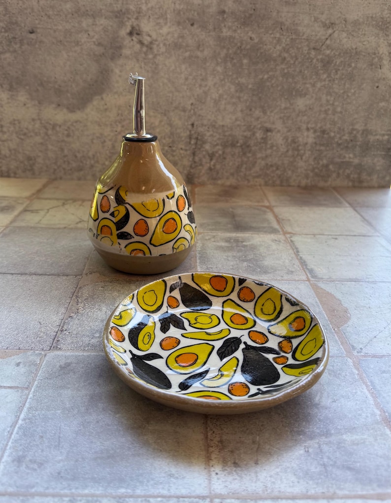 May include: A ceramic oil bottle and small dish set. The bottle has a brown base and a silver spout, decorated with yellow, orange, and black avocado designs. The matching dish features the same avocado pattern. A unique kitchenware set.
