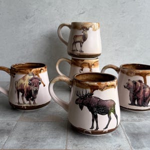 May include: A collection of ceramic mugs featuring wildlife illustrations. Each mug has a cream-colored base with a brown rim and handle. The mugs depict a bison, moose, bear, and deer. The mugs are stacked and arranged on a tiled surface.