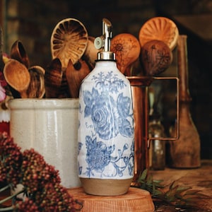 May include: A ceramic oil dispenser featuring a white base with a blue floral pattern and a metallic pour spout. The cylindrical vessel has a smooth finish on the upper portion and a natural, unglazed tan base, standing upright.