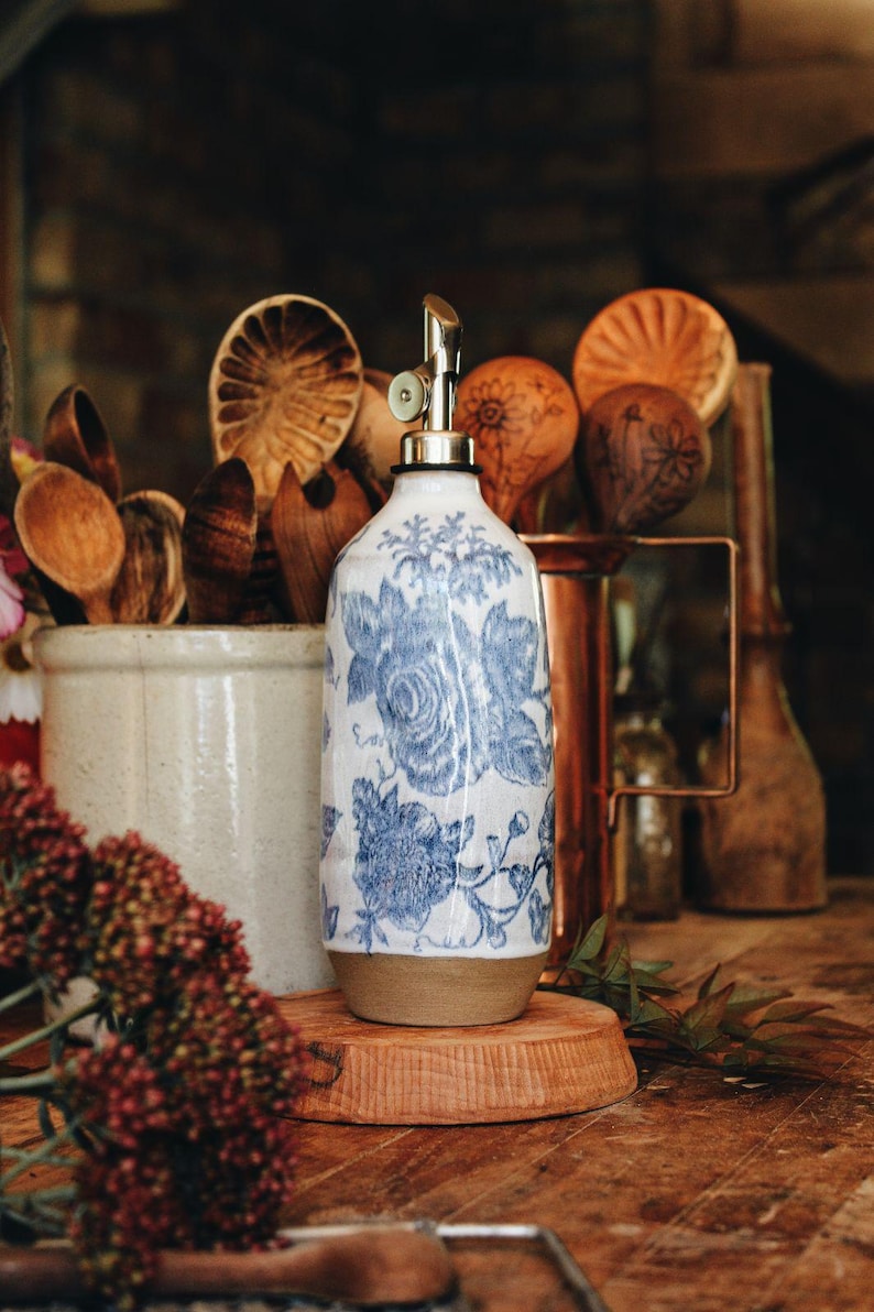 May include: A ceramic oil dispenser featuring a white base with a blue floral pattern and a metallic pour spout. The cylindrical vessel has a smooth finish on the upper portion and a natural, unglazed tan base, standing upright.
