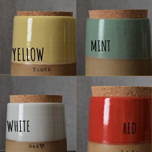 Op de afbeelding: Vier keramische potten met kurken deksels, elk in een andere kleur: geel, mint, wit en rood. Het woord voor elke kleur staat op de pot. De onderste helft van elke pot is een natuurlijke bruine kleur.