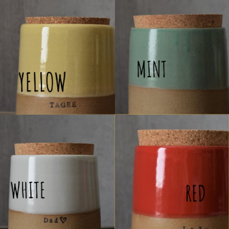 Op de afbeelding: Vier keramische potten met kurken deksels, elk in een andere kleur: geel, mint, wit en rood. Het woord voor elke kleur staat op de pot. De onderste helft van elke pot is een natuurlijke bruine kleur.